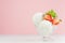 Three creamy ice cream balls in elegant glass bowl with green mint, strawberry pieces on white wood table and pastel pink wall.