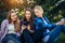 Three cute girls relax and socialize on the lawn in summer park. Young women sit on the green grass among the trees and look