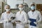 Three factory workers wearing lab coats and masks looking at camera in workshop