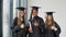 Three female students of different races with a diploma in hand. Graduates in black robes and square hats look at the