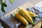 Three fresh corn cobs with leaves on a wooden table over grey wall background. Raw corncobs on light brown wooden table, rustic