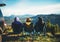 Three friends sit in camping chairs on top of sun mountain, travelers enjoy nature, tourists look into distance on background