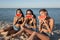 Three funny girls in jean shorts and black bras are sitting on the sand near the sea and holding watermelon slices near