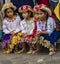 Three girls in traditional Ecuadorian clothes sit on street curb watching parade