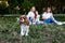 Three girls, wearing white shirts and blue jeans, sitting on grass, playing with small dog in park. Cavalier king charles spaniel