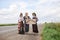 Three hippie women, wearing boho style clothes, standing on road, thumbing a ride, hitchhiking with sign Happy place on cardboard