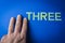 Three human fingers beside the word Three written with plastic letters on blue paper background