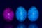 Three marbled, colored, blue and pink Easter eggs stand side by side against a reflective blue background and are reflected on the