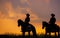 Three men dressed in cowboy garb, with horses and guns. A cowboy riding a horse in the sunset is silhouetted in black