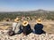 Three Mexican male tourists in straw hats sit with their backs to the camera on top of the pyramid against the backdrop of mountai