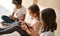 Three multiethnic little kids sit indoor using digital gadgets