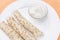 Three pancakes roll with stuffing and sour cream in a bowl in a white plate on a delicate peach-orange background, close-up