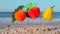 Three paper fruits on stick stand on sandy beach near sea on sunny summer day.