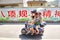 Three person family rides a scooter with umbrella protecting from the sun.