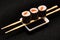 Three pieces of syake maki roll lying on sticks on a black background.