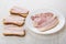 Three sandwiches with bacon, plate with slices of bacon on table