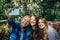 Three sisters take a selfie on smartphone sitting in a park on a background of flowers on sunny day. Brunette, blonde and redhead