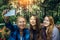 Three sisters take a selfie on smartphone sitting in a park on a background of flowers on sunny day. Brunette, blonde and redhead