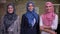 Three smiling arab females wearing hijab, pink, blue and grey are standing and looking at camera, in modern brick office