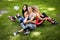Three sportive female friends sitting on a grass at the park outdoors, make selfie on a phone with a stick