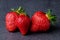 Three strawberries in a row on dark grey background with green leaves, ripe fruits, macro shot made in a professional studio,