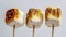 Three Toasted Marshmallows On Sticks With Varying Degrees Of Charring Against A Light Background