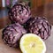 Three Tuscan artichokes and a slice of lemon lit by sunlight on a wooden chopping board