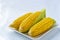 Three Vibrant Yellow Corn Cobs With Green Husks On White Plate, Set Against A Soft Blue Background