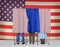 Three Voting booths in front of a large American Flag. Two Women and one man behind curtains one woman is starting to leave.