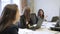 Three women listening and talking while sitting at table with laptops in office.