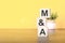 Three wooden cubes with letters - M and A, on yellow background, space for text in left. MA - short for Mergers and