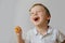 A three-year-old boy laughs after biting off a delicious orange fruit. The boy is wearing a white T-shirt