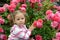The three-year-old thoughtful girl against the background of a bush of the blossoming roses