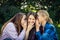 Three young attractive woman sharing secrets sitting on green grass in the park. Cheerful girlfriends gossip and whisper outdoor