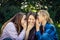 Three young attractive woman sharing secrets sitting on green grass in the park. Cheerful girlfriends gossip and whisper outdoor