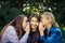 Three young attractive woman sharing secrets sitting on green grass in the park. Cheerful girlfriends gossip and whisper outdoor