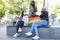 Three young caucasian and asian homosexual friends sitting on a LGBT flag bench wearing face masks.