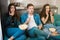 Three young friends two beautiful women and handsome man with remote control watching scary scene in horror movie on the sofa