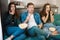 Three young friends two beautiful women and handsome man with remote control watching scary scene in movie sit on the sofa eating