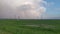 A thundercloud is moving over a sown field with power line poles standing on it. It is raining and a rainbow is visible.