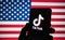 TikTok logo seen on the silhouette of smartphone hold in a hand with blurred American flag on tha back. Rela photo, not a montag