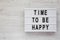 `Time to be happy` words on a lightbox on a white wooden surface, top view. Overhead, from above. Flat lay. Copy space