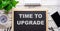 TIME TO UPGRADE is written in gray on a black background next to a white alarm clock, a green plant and a notepad. Motivational