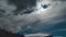 A timelapse of black and gray clouds moving against a full moon and stars. A black silhouette of a mountain and trees in