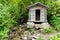 Tiny wooden mountain cabin in dense lush green summer forest with a small creek beside it