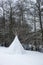 Tipi tent covered with snow next to a river by the forest. Winter glamping outdoors, camp site