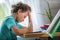 Tired boy puts his hand on head and looks away, sitting at Desk and doing lesson