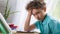 Tired boy puts his hand on head and looks away, sitting at Desk and doing lesson