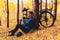 Tired cyclist is sitting on fall leaves in forest and resting. Togetherness with nature