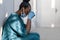 Tired depressed female african nurse wearing face mask sits on hospital floor.